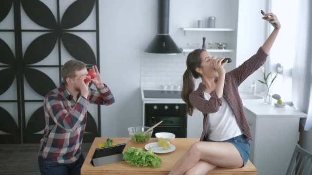 crazy family, funny cheerful man is fooling around and making grimaces with vegetables and woman takes picture on smartphone while sitting on table in kitchen while cooking healthy food for lunch