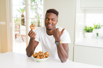 African american man eating chocolate chips muffin pointing and showing with thumb up to the side with happy face smiling
