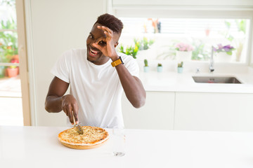 African american man eating cheese pizza at home with happy face smiling doing ok sign with hand on eye looking through fingers