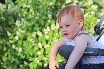 little, beautiful, smiling, cute redhead baby in a sleeveless shirt in a pram out-of-doors drops hands down and looking forward