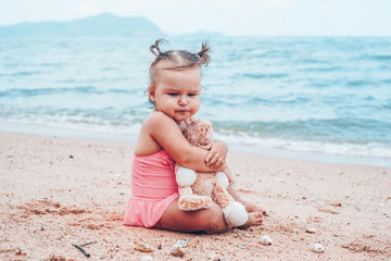 pensive little girl hugging teddy bear and looking away while sitting on seashore.
