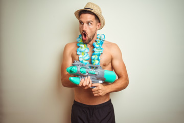 Handsome shirtless man wearing hawaiian lei and water gun over background scared in shock with a surprise face, afraid and excited with fear expression