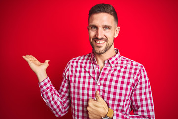 Fototapeta premium Young handsome man over red isolated background Showing palm hand and doing ok gesture with thumbs up, smiling happy and cheerful
