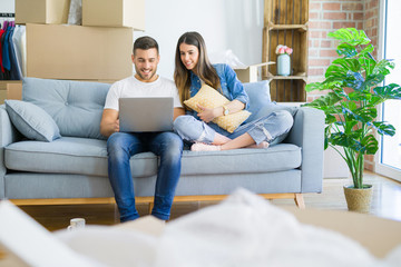 Obraz premium Young beautiful couple relaxing sitting on the sofa of new house using computer laptop with a happy face standing and smiling with a confident smile showing teeth