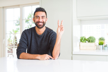 Handsome hispanic man wearing casual sweater at home smiling with happy face winking at the camera doing victory sign. Number two.