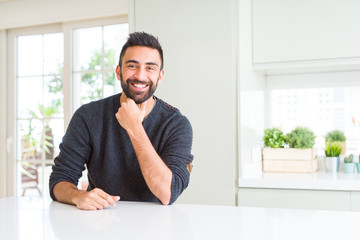 Fototapeta premium Handsome hispanic man wearing casual sweater at home looking confident at the camera with smile with crossed arms and hand raised on chin. Thinking positive.