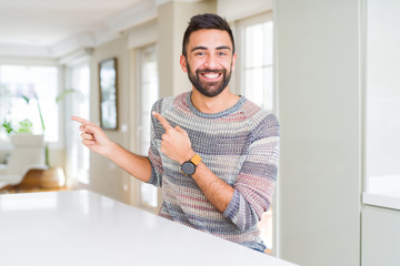 Handsome hispanic man wearing casual sweater at home smiling and looking at the camera pointing with two hands and fingers to the side.