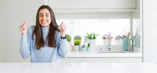 Wide angle picture of beautiful young woman sitting on white table at home excited for success with arms raised celebrating victory smiling. Winner concept.