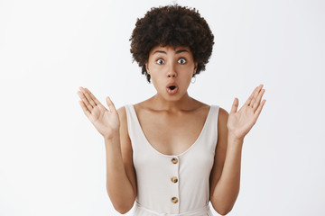 Indoor shot of attractive african american female in stylish outfit with curly hairstyle raising palms and folding lips popping eyes from amazement and curiousity, being intrigued by fresh rumors