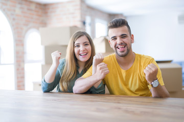 Young couple sitting on the table movinto to new home with carboard boxes behind them celebrating surprised and amazed for success with arms raised and open eyes. Winner concept.