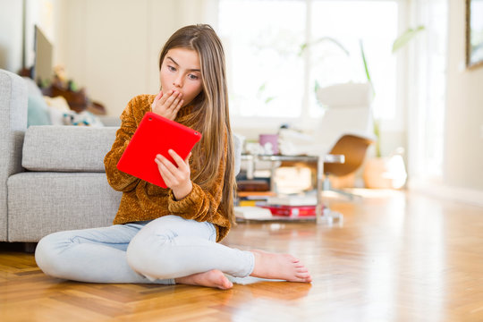 Beautiful young girl kid using digital touchpad tablet sitting on the floor cover mouth with hand shocked with shame for mistake, expression of fear, scared in silence, secret concept