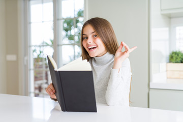 Beautiful young girl reading a book at home very happy pointing with hand and finger to the side