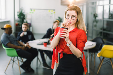 Cheerful young beautiful blond woman in glasses and red shirt standing at her working place on office with cup of coffee. Multiracial young creative people in modern office.