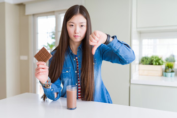 Beautiful Asian woman drinking chocolate milkshake and holding chocolate bar with angry face,...