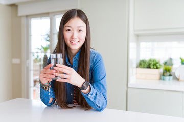 Beautiful Asian woman drinking a glass of fresh water with a happy face standing and smiling with a confident smile showing teeth