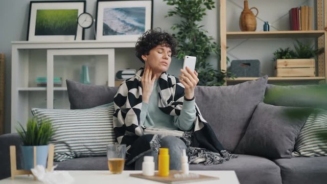 Young lady patient is skyping talking to doctor using smartphone discussing treatment for disease sitting on sofa in apartment. Millennials and technology concept.