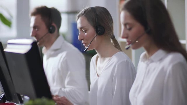 Selective focus on smiling attractive young woman customer support operator talking via headset and actively typing the keyboard of desktop computer. Working hard, profession concept, career. Female
