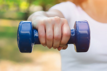 Sport training concept. Female hand is holding a blue dumbbell against the background of the park. The girl is engaged in fitness in nature