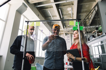 Three multiethnical businesspeople in casual wear discussing and planning concept, working together in loft office with glass wall. Front of glass wall marker and stickers. Startup office.