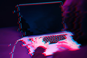 Hacker in the hood working with laptop typing text in dark room. Image with glitch effect