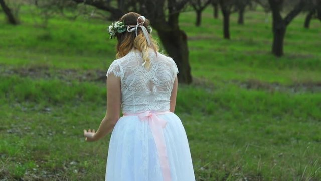 Ballerina in white wedding dress poses with spread out hands in apple orchard among trees in ballerinas outdoors on grass during spring day closeup view captured in 120 FPS
