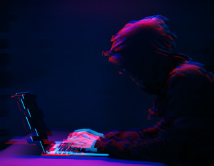 Hacker in the hood working with laptop typing text in dark room. Image with glitch effect
