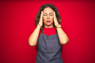 Middle age senior woman wearing apron uniform over red isolated background with hand on headache because stress. Suffering migraine.