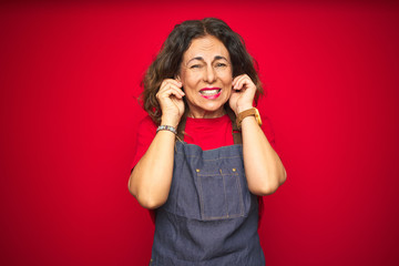 Middle age senior woman wearing apron uniform over red isolated background covering ears with fingers with annoyed expression for the noise of loud music. Deaf concept.