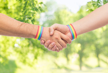 lgbt, same-sex love and homosexual relationships concept - close up of male couple hands with gay pride rainbow awareness wristbands making handshake over green natural background