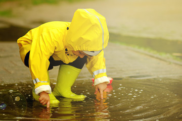 Cute child playing in a puddle with a ship on a summer rainy day. a boy in a yellow raincoat walks in the park. - obrazy, fototapety, plakaty