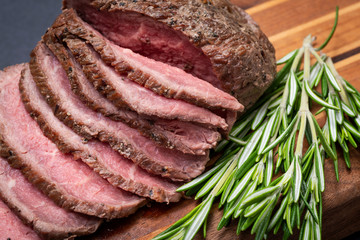 Sliced Grass Fed juicy Corn Roast Beef garnished with Rosemary Fresh Herb on natural wood cutting board.