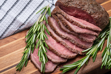 Sliced Grass Fed juicy Corn Roast Beef garnished with Rosemary Fresh Herb on natural wood cutting board.