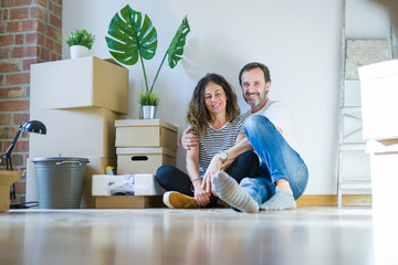 Middle age senior romantic couple in love sitting on the apartment floor with cardboard boxes around and smiling happy for moving to a new home
