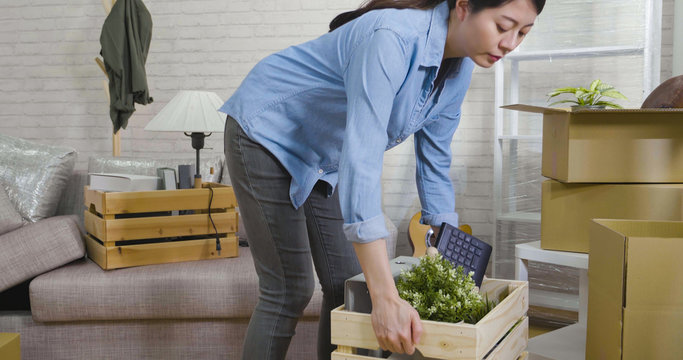 Beautiful Asian Japanese Young Girl Moving In New Apartment Standing In Living Room Put Down Wood Box Full Of Stuff On Floor. Independent Female Carry Heaving Container With Belongings Inside Home