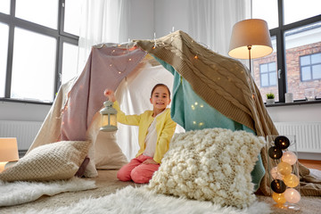 childhood and hygge concept - happy little girl with lantern in kids tent or teepee at home