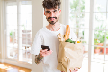 Young handsome man holding a paper bag full of fresh groceries at home, showing and using smartphone while buying products