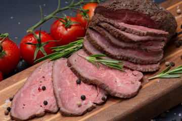 Sliced Grass Fed juicy Corn Roast Beef garnished with Tomatoes, Fresh Rosemary Herb and Rainbow Peppercorns on natural wooden cutting board.