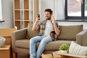 Obraz premium moving, people and real estate concept - happy man with smartphone and boxes sitting on sofa and having video call at new home