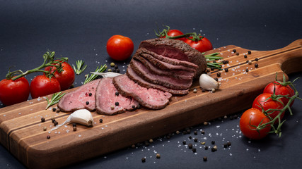 Sliced Grass Fed juicy Corn Roast Beef garnished with Tomatoes, Fresh Rosemary Herb, Garlic and Rainbow Peppercorns on natural wooden cutting board.