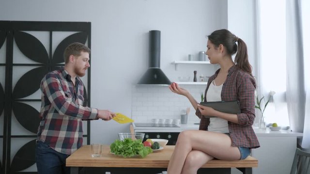 useful food, happy guy prepares healthy vegetarian meal on dinner and girl sitting on kitchen table with tablet in hands and communicate at cuisine