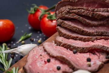 Sliced Grass Fed juicy Corn Roast Beef garnished with Tomatoes, Fresh Rosemary Herb, Garlic and Rainbow Peppercorns on natural wooden cutting board.