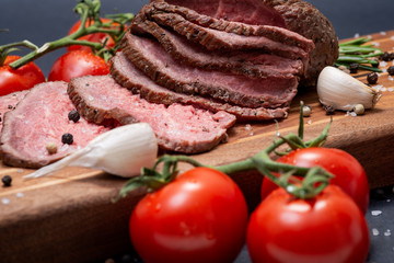 Sliced Grass Fed juicy Corn Roast Beef garnished with Tomatoes, Fresh Rosemary Herb, Garlic and Rainbow Peppercorns on natural wooden cutting board.