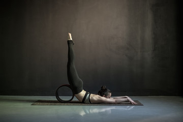 Woman practicing yoga, doing Salabhasana pose with wheel. Indoor full length, black wall yoga studio