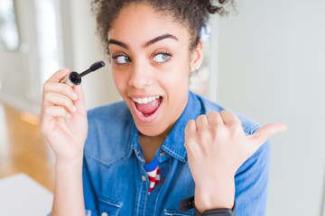 Young african american girl applying eyelashes mascara pointing and showing with thumb up to the side with happy face smiling