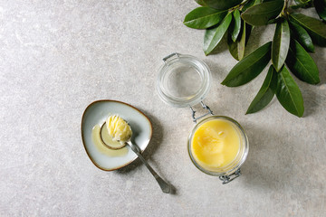 Homemade Melted ghee clarified butter in open glass jar and spoon on saucer over grey texture background with green branch. Flat lay, space