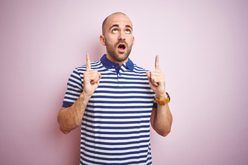 Young bald man with beard wearing casual striped blue t-shirt over pink isolated background amazed and surprised looking up and pointing with fingers and raised arms.