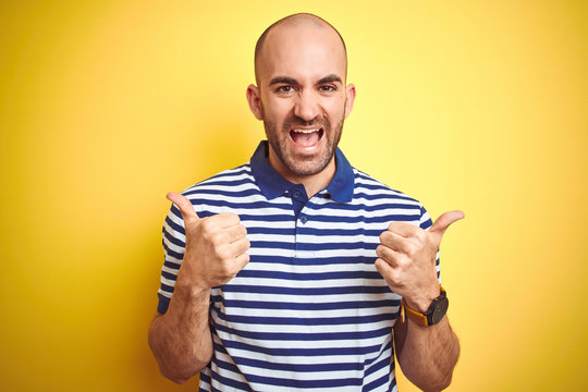 Young bald man with beard wearing casual striped blue t-shirt over yellow isolated background success sign doing positive gesture with hand, thumbs up smiling and happy. Cheerful expression and winner