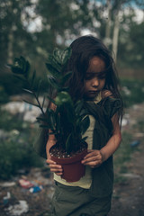 A homeless girl is standing on a garbage dump with a houseplant in a pot.