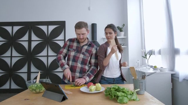young funny woman with man dancing and having fun while cooking healthy eating from vegetables on lunch for wellness according to diet plan on cuisine table