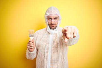 Young man wearing painter equipment and holding painting brush over isolated yellow background pointing with finger to the camera and to you, hand sign, positive and confident gesture from the front